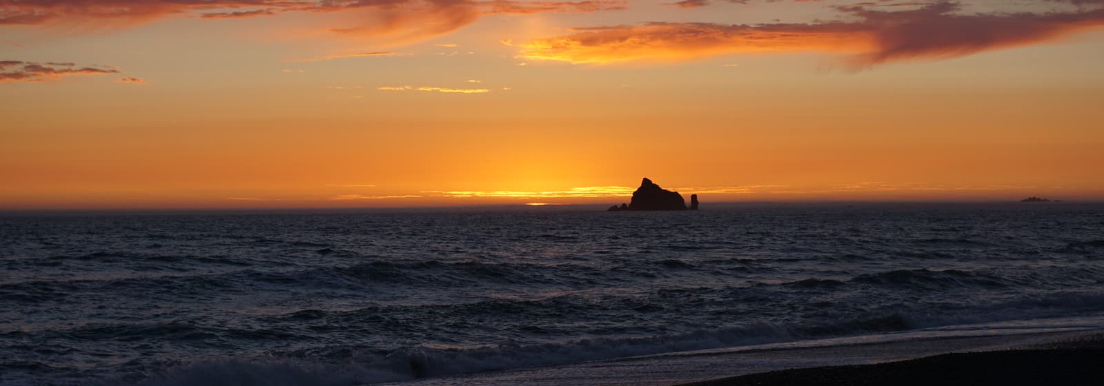 A sea stack silhouetted against an orange sunset over the Pacific, waves rolling toward a dark gravel beach